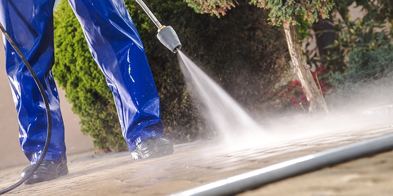 Two individuals are cleaning a spacious area with a vacuum and mop, while another uses a pressure washer nearby. They are all wearing matching uniforms and gloves, with various cleaning equipment and plants visible around the office setting.